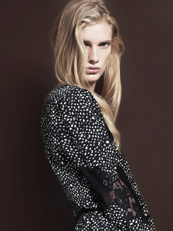 Portrait of a blonde adult woman in a patterned black and white dress with lace sleeves, posing against a solid brown backdrop.