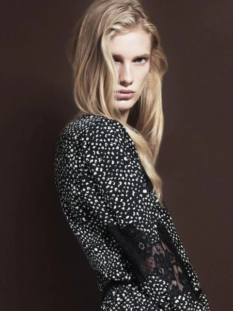 Portrait of a blonde adult woman in a patterned black and white dress with lace sleeves, posing against a solid brown backdrop.