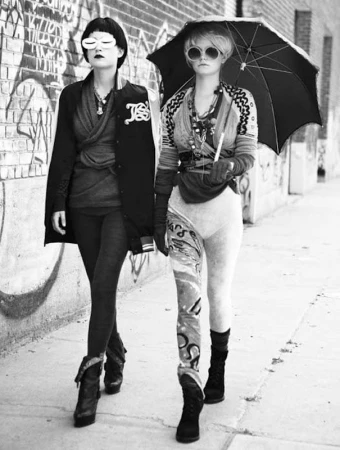 Two women walking side by side down an urban street, one holding a black umbrella and wearing patterned leggings.