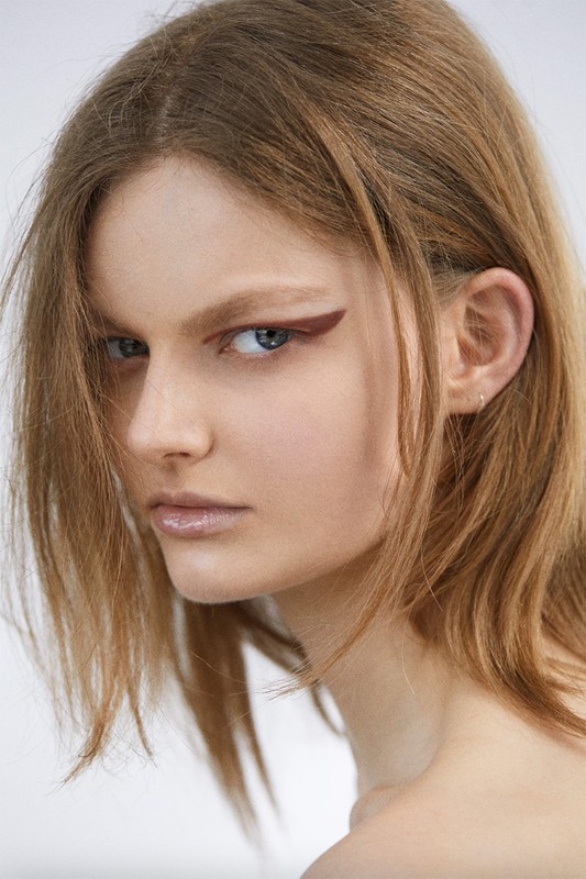 Close-up portrait of a young woman with brown hair and sharp graphic eyeliner, shot against a plain light background