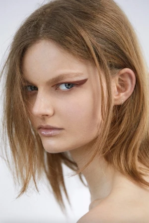 Close-up portrait of a young woman with brown hair and sharp graphic eyeliner, shot against a plain light background