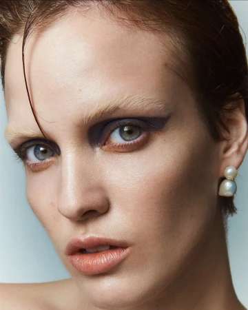 Close-up portrait of a model with short wet-look hair, dramatic purple eyeshadow, and pearl earrings against a light background