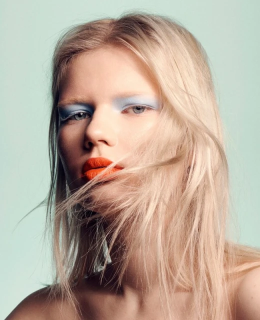 Close-up portrait of a young adult woman with orange lipstick, blue eyeshadow, and flowing blonde hair against a light blue background.