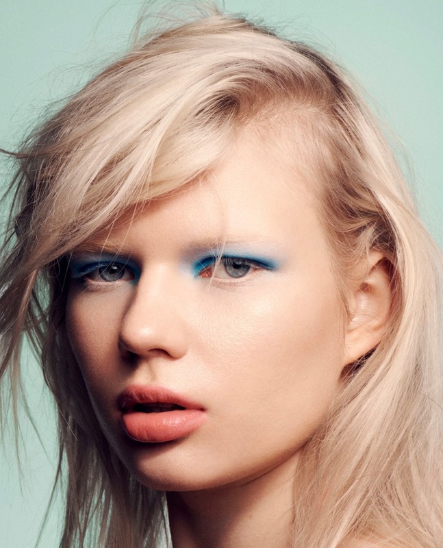 Close-up portrait of a blonde woman with blue eye makeup, fair skin, and pink lips against a light blue background in a beauty shot.