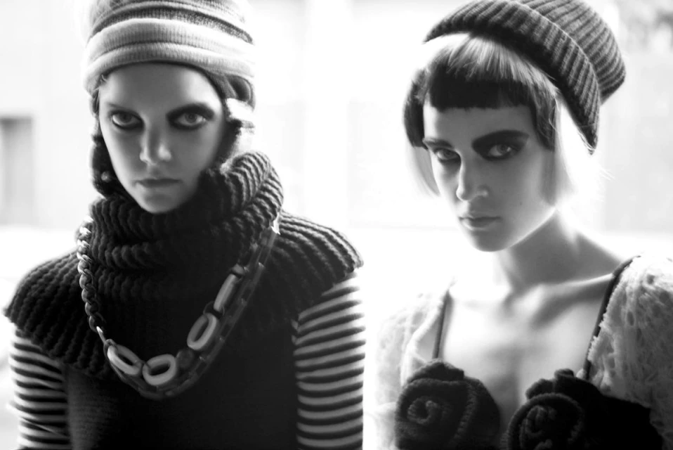 Two fashion models pose in knit hats and tops against a bright background, showcasing a monochrome editorial style with dark makeup.