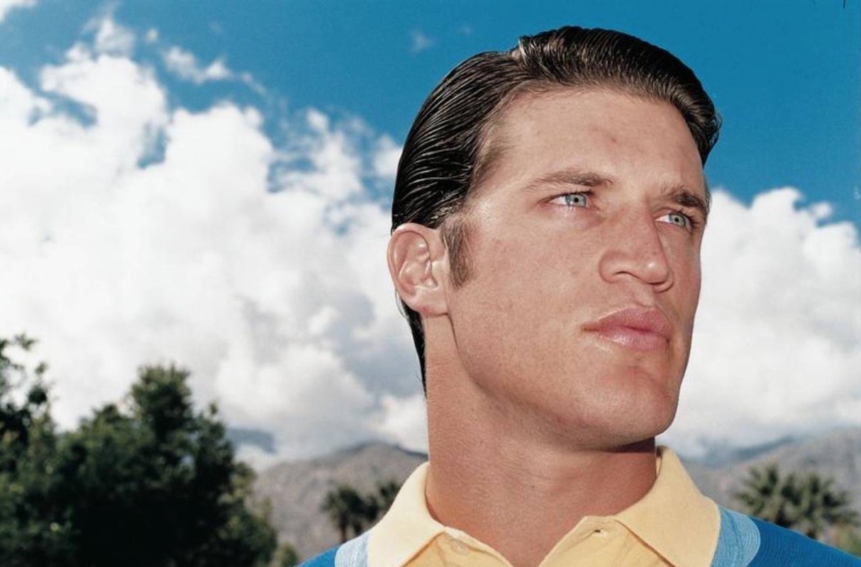 Headshot of an adult male with slicked-back brown hair and blue eyes, wearing a yellow polo shirt, set against a background of blue sky and clouds.