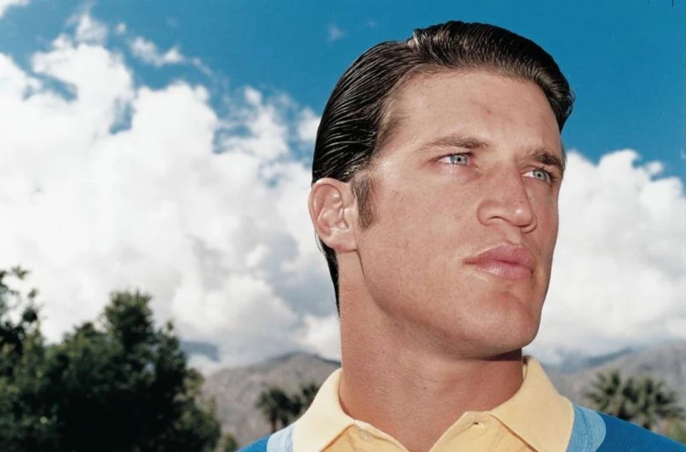 Headshot of an adult male with slicked-back brown hair and blue eyes, wearing a yellow polo shirt, set against a background of blue sky and clouds.