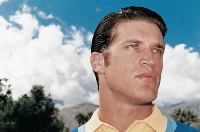 Headshot of an adult male with slicked-back brown hair and blue eyes, wearing a yellow polo shirt, set against a background of blue sky and clouds.