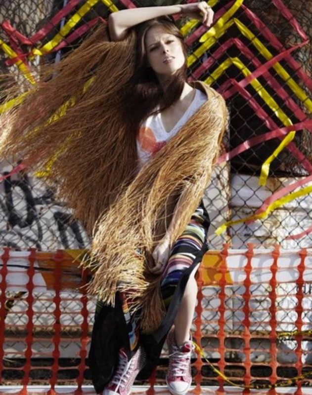 A stylish woman in a faux fur shawl and Converse stands in front of a safety fence with ribbons, creating an urban fashion portrait.