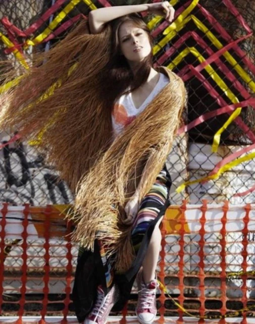 A stylish woman in a faux fur shawl and Converse stands in front of a safety fence with ribbons, creating an urban fashion portrait.