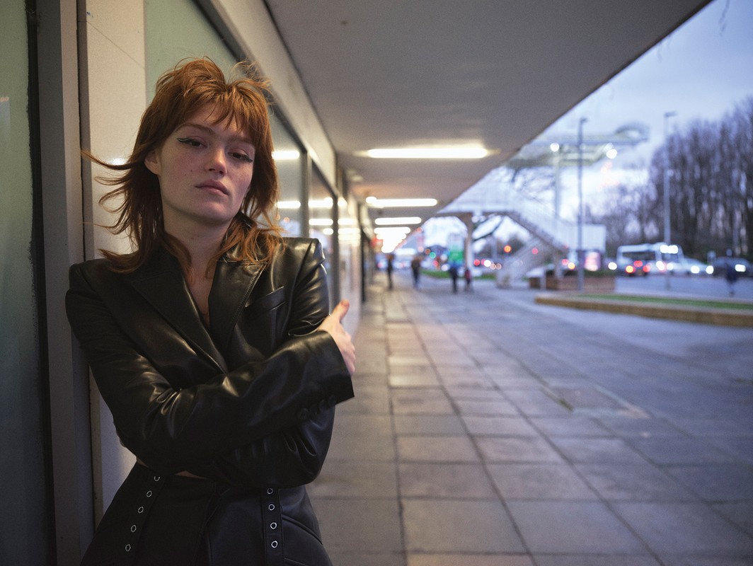 A young woman with reddish hair wearing a black leather jacket is posing outdoors in an urban setting near a tiled walkway.