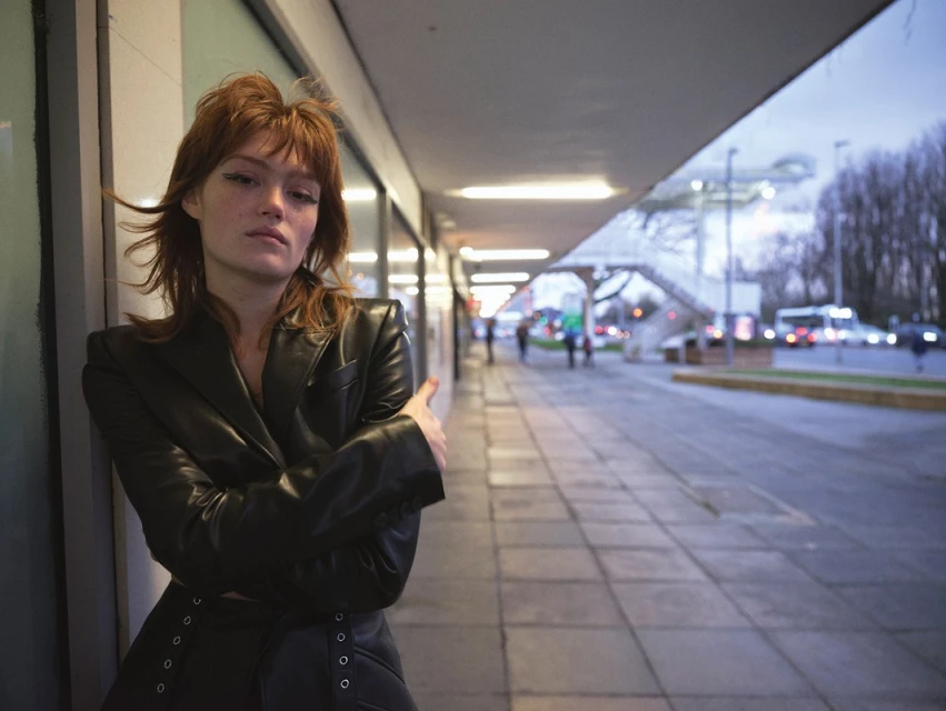 A young woman with reddish hair wearing a black leather jacket is posing outdoors in an urban setting near a tiled walkway.