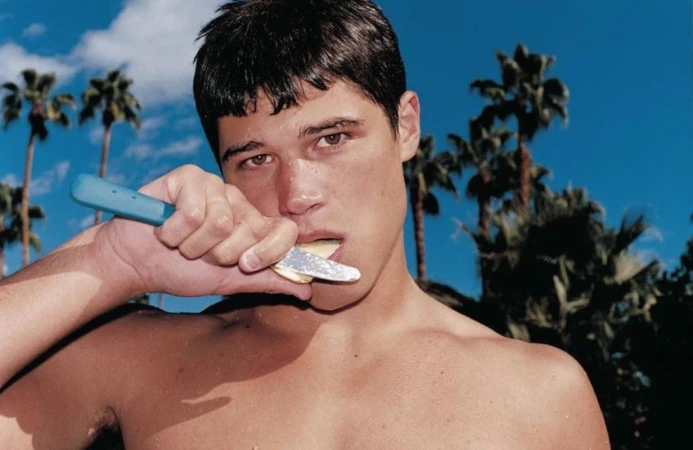 Shirtless man holding a blue-handled knife to his mouth against a bright blue sky and palm trees