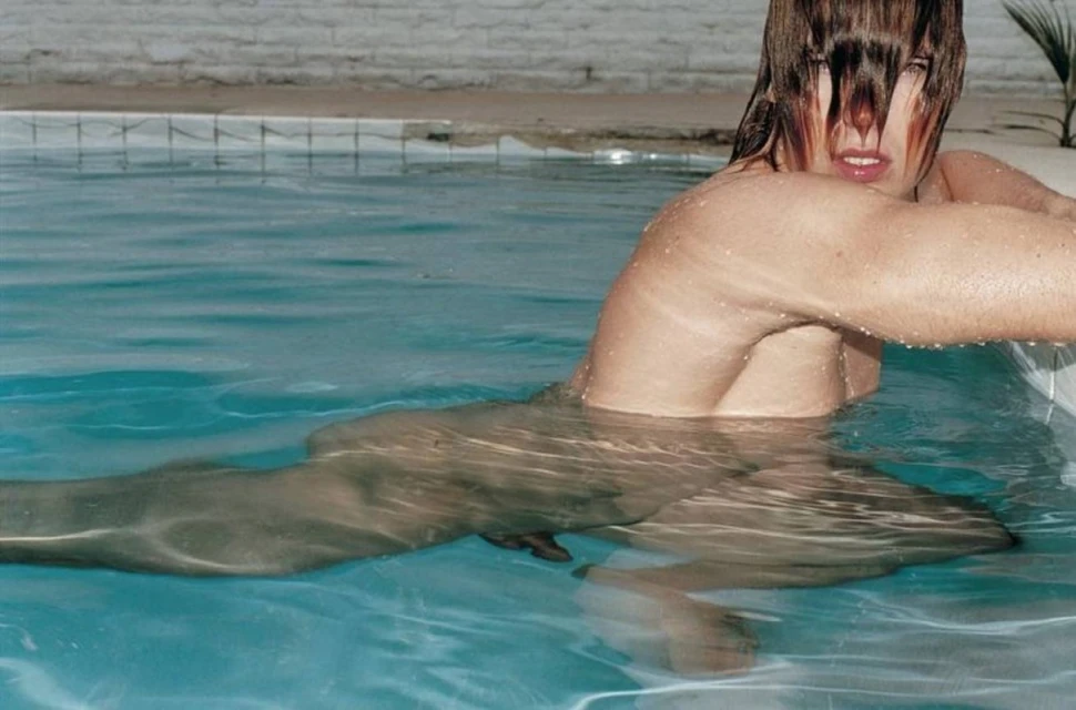 Adult female partially submerged in a turquoise-blue swimming pool, showcasing wet blonde hair, fair skin, and water droplets.
