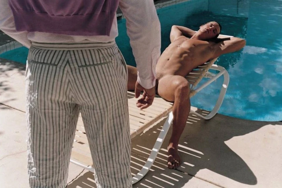 Nude man reclines on chair by turquoise pool, juxtaposed with striped-trousered individual, sunny outdoor scene with light shadows.