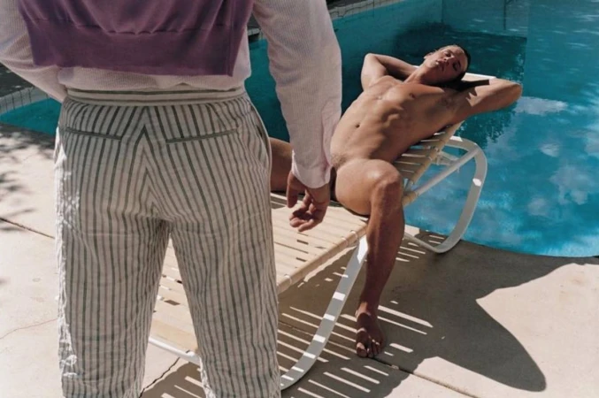 Nude man reclines on chair by turquoise pool, juxtaposed with striped-trousered individual, sunny outdoor scene with light shadows.