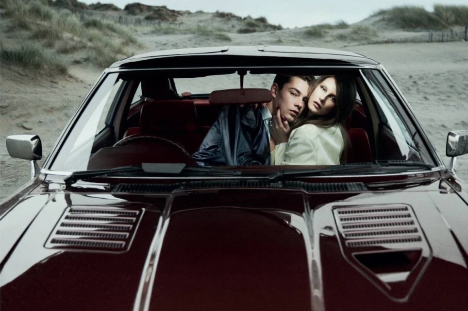 Automotive fashion shoot featuring a couple in a vintage car against a beach backdrop with grassy dunes and overcast sky, stylish attire.