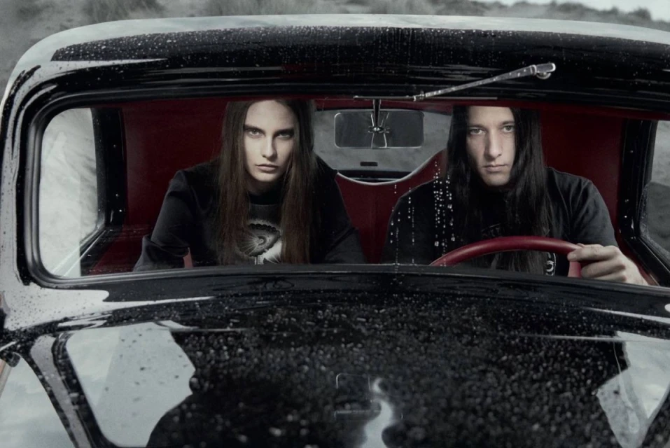 Two people in the front seats of a black vintage car, one driving and the other in the passenger seat, looking out.