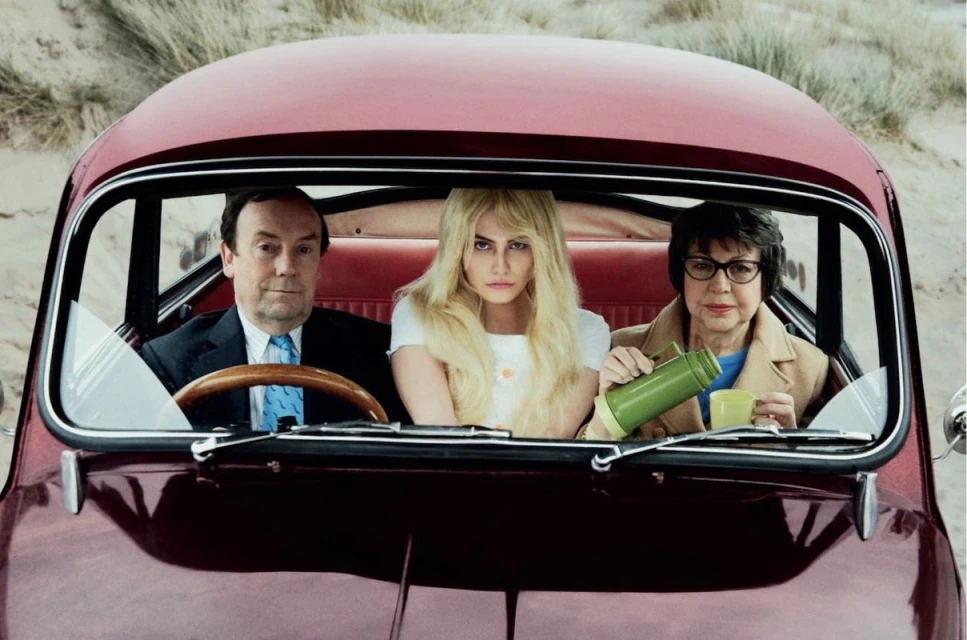 Three people in vintage car: a man in a suit, a young blonde woman, and an elderly woman with a thermos against a neutral backdrop.