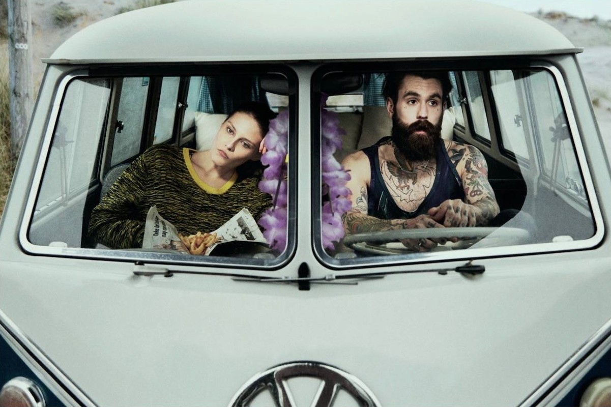 Woman and man sit in a vintage Volkswagen van on a beach. The woman reads a newspaper while the man looks ahead, both with neutral expressions.