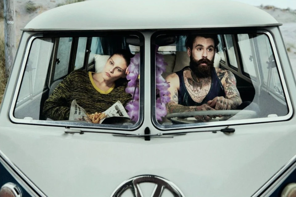Woman and man sit in a vintage Volkswagen van on a beach. The woman reads a newspaper while the man looks ahead, both with neutral expressions.
