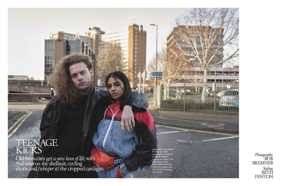Two young people, a man and a woman, stand close together on an urban street, dressed in casual outerwear, with buildings in the background.