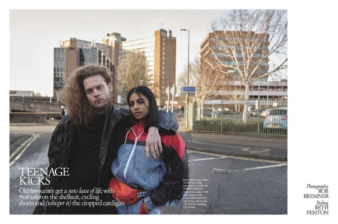 Two young people, a man and a woman, stand close together on an urban street, dressed in casual outerwear, with buildings in the background.