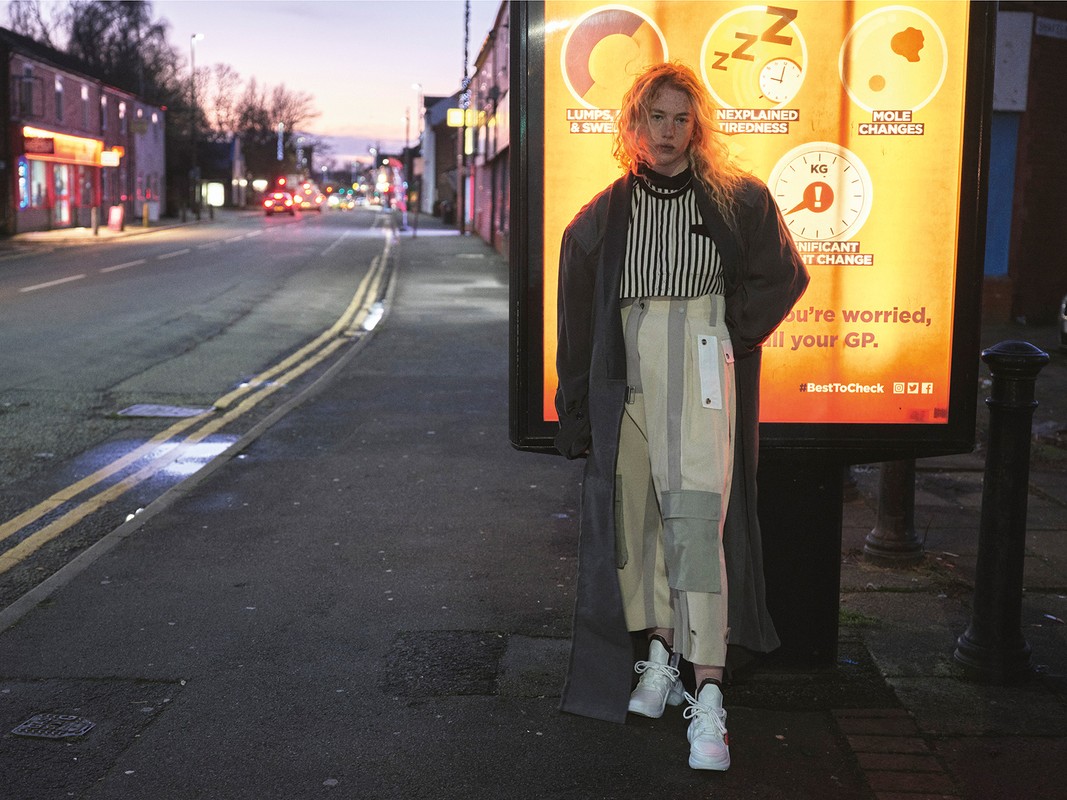 Woman stands on a city sidewalk at dusk, wearing striped top, high-waisted pants, sneakers, and a coat; street and buildings in background.