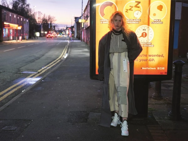 Model with blonde curly hair wearing a long grey coat and striped top standing on a street next to an illuminated billboard