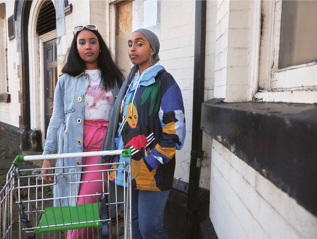 Two women pose with a shopping cart in front of a weathered building, showcasing casual style and unique makeup in an urban setting.
