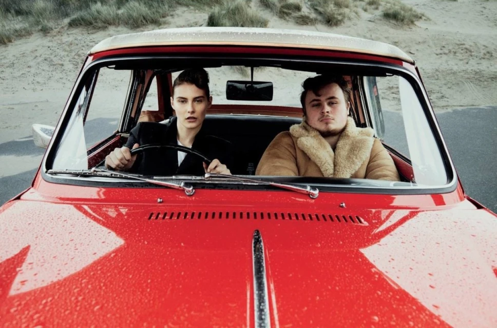 A stylish woman driving a vintage red car with a man in the passenger seat on a cloudy day by a sandy beach, capturing a vintage fashion moment.