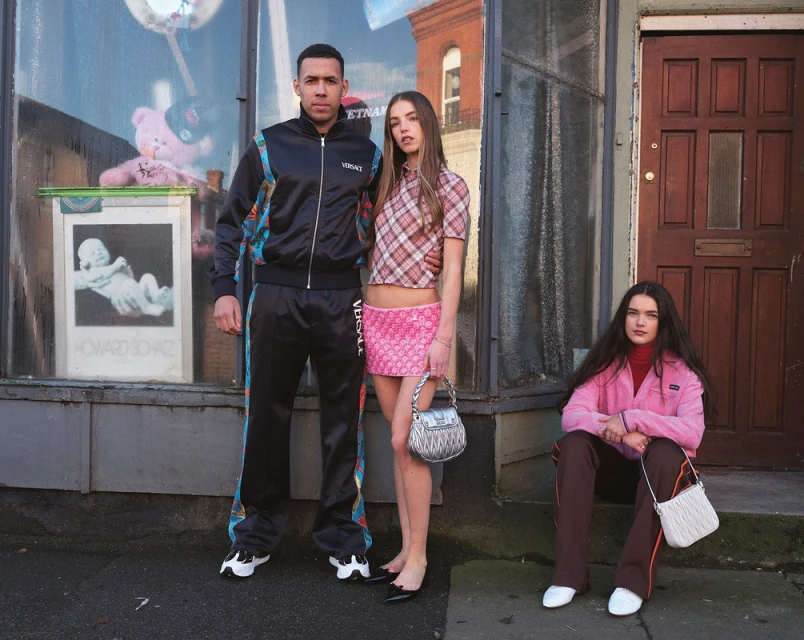Three young adults posing outside a brick building. One man in tracksuit, two women in skirts and jackets, overcast daylight.