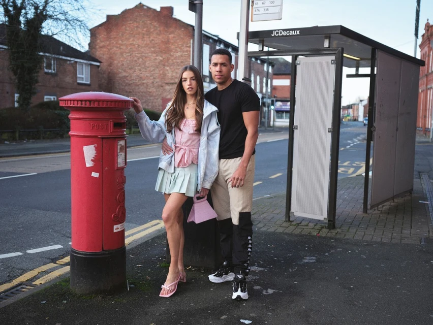 Young adult couple poses next to a red British telephone box in an urban environment. The woman is holding a small purse.