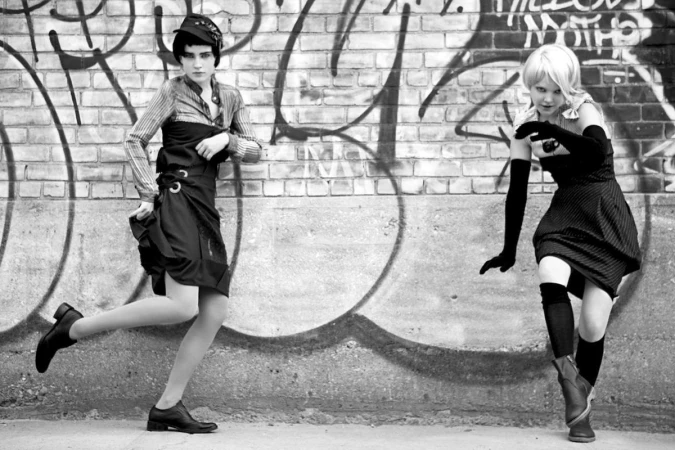 Two women posing playfully in front of a weathered brick wall covered in graffiti tags.