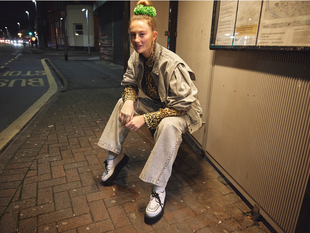Young woman in a denim jacket sits at a bus stop at night, illuminated by street lights, creating a casual, urban fashion portrait.