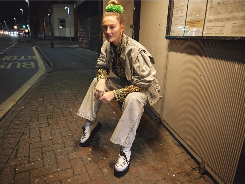 Young woman in a denim jacket sits at a bus stop at night, illuminated by street lights, creating a casual, urban fashion portrait.
