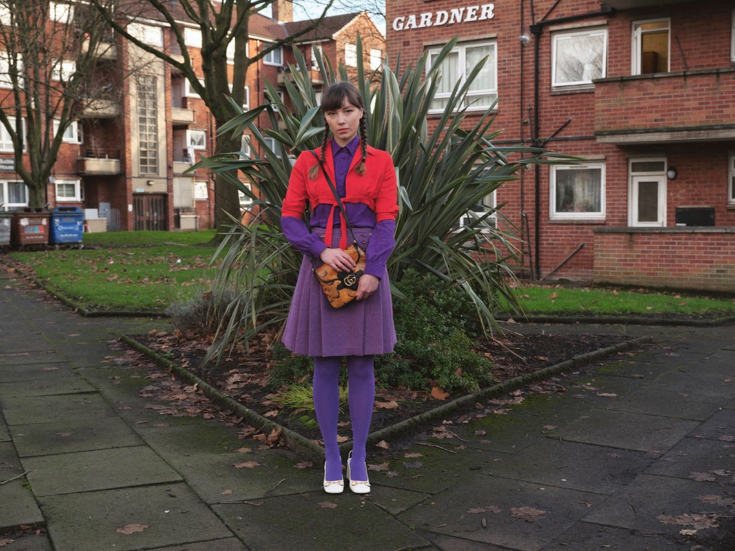 Woman in a purple skirt, tights, and red jacket stands outside a red brick building with shrubbery and a sidewalk in an urban setting.