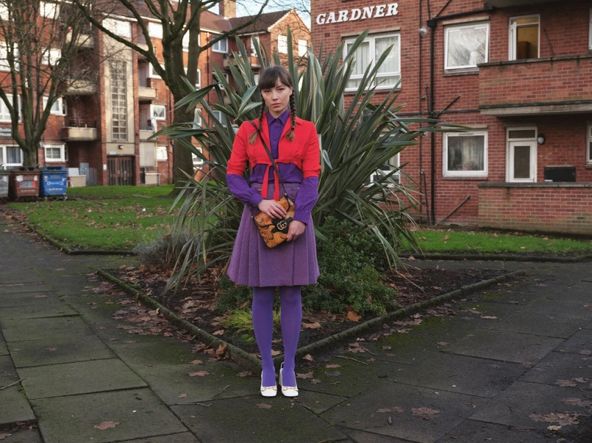 Woman in a purple skirt, tights, and red jacket stands outside a red brick building with shrubbery and a sidewalk in an urban setting.