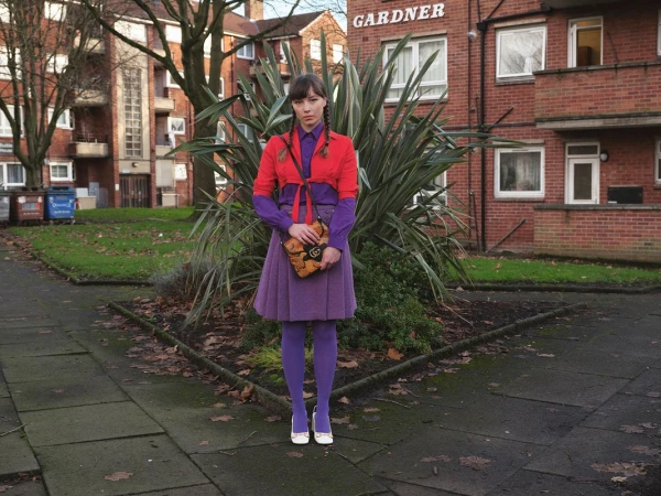 Woman in a purple skirt, tights, and red jacket stands outside a red brick building with shrubbery and a sidewalk in an urban setting.