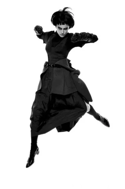 Full-length black and white shot of a female fashion model leaping mid-air in an all-black avant-garde outfit against a white backdrop.