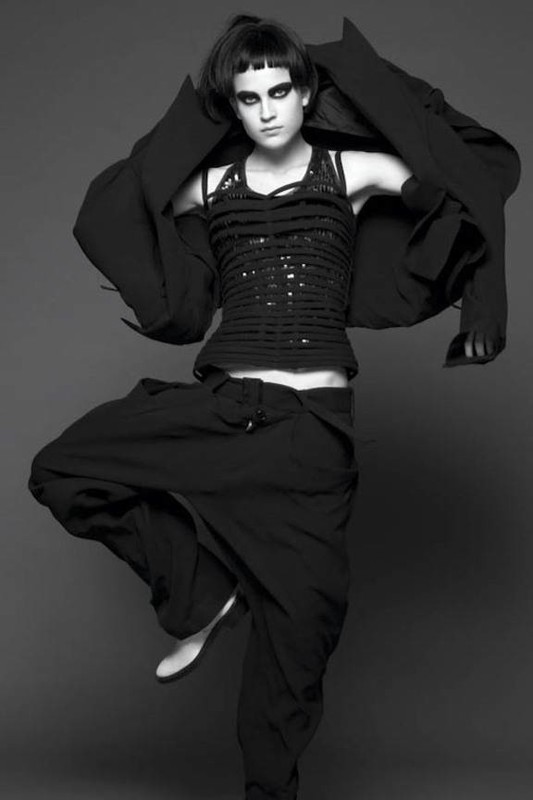 Full-length black and white portrait of a woman in black clothing striking a dance pose on a gray background in a studio setting.