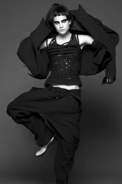 Full-length black and white portrait of a woman in black clothing striking a dance pose on a gray background in a studio setting.