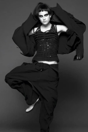 Full-length black and white portrait of a woman in black clothing striking a dance pose on a gray background in a studio setting.