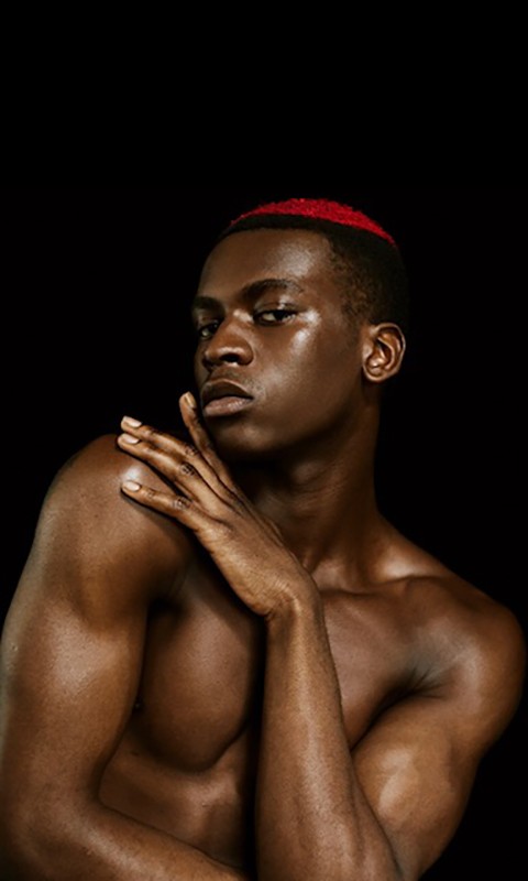 Striking portrait of shirtless Black male model with red hair accent against black backdrop, showcasing skincare, beauty, and fashion.