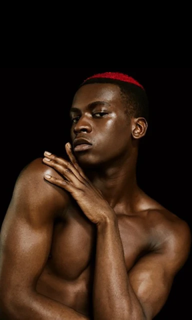 Striking portrait of shirtless Black male model with red hair accent against black backdrop, showcasing skincare, beauty, and fashion.