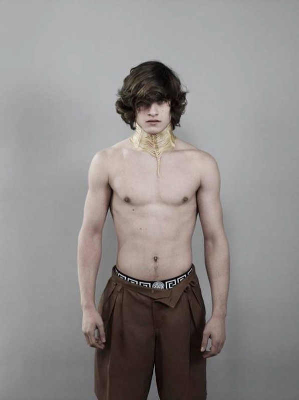Portrait of a shirtless adult male with brown hair and dark brown pleated dress shorts against a neutral gray background in a photo studio.