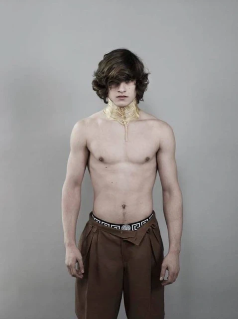 Portrait of a shirtless adult male with brown hair and dark brown pleated dress shorts against a neutral gray background in a photo studio.