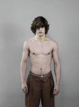 Portrait of a shirtless adult male with brown hair and dark brown pleated dress shorts against a neutral gray background in a photo studio.