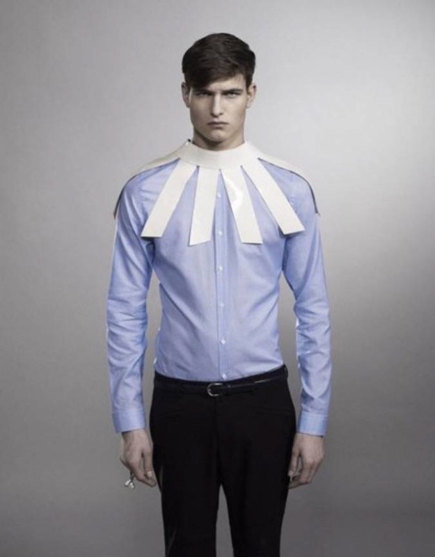 Fashion shot of a young adult male in a light blue shirt and paper collar standing against a gray background in a studio setting.