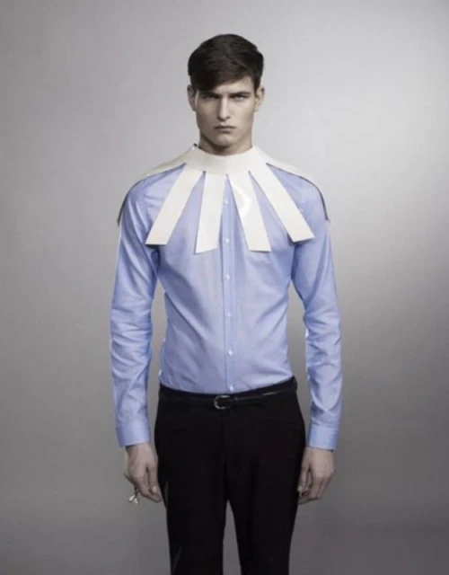 Fashion shot of a young adult male in a light blue shirt and paper collar standing against a gray background in a studio setting.